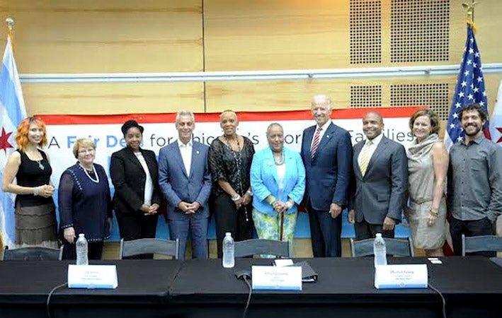 Entrepreneur and chainmaille expert Rebeca Mojica standing (far left, with orange hair and a black top) in a professional group photo with former US Vice President Joe Biden (7th from right), former Chicago Mayor Rahm Emanuel (4th from left), and seven other business and policy leaders at a small business roundtable event. The US and Chicago flags are visible in the corners.