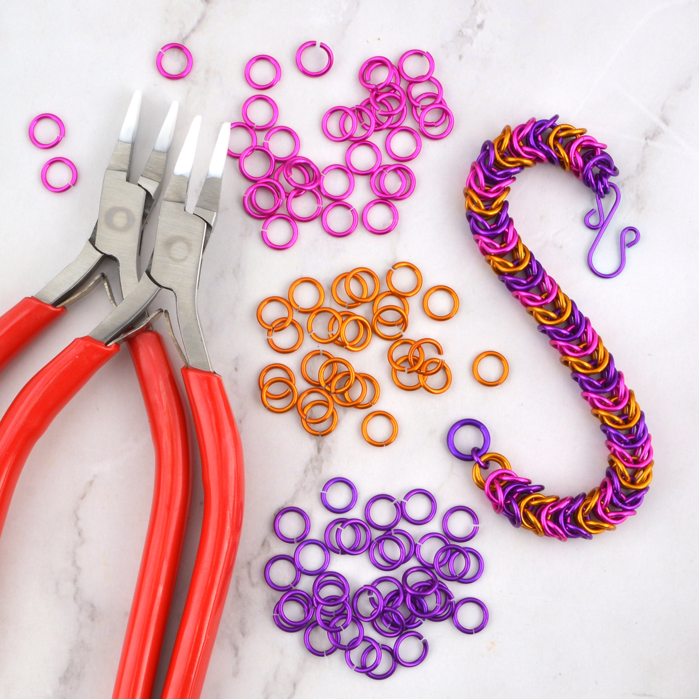 A flat-lay of DIY chainmaille materials including red-handled pliers, loose purple, pink, and orange jump rings, and a finished slinky-box weave bracelet in coordinating colors.