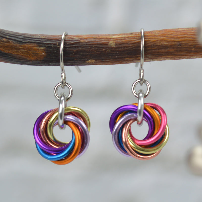 Tiny chainmaille earrings hanging from a branch. Each earring has a small "knot" of 7 jump rings to best match the colors of the xenogender pride flag: pink, peach, orange, pastel yellow, azure, lilac, and violet. Earrings are attached to stainless steel earwire. The background is a blurry grey brick wall.