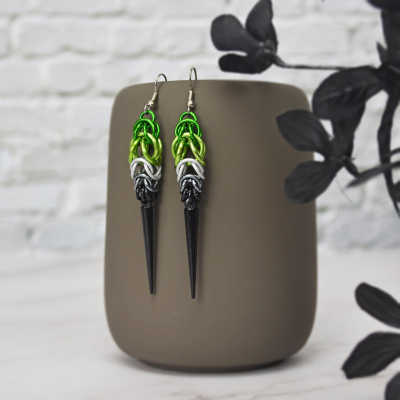 Aromantic pride chainmaille earrings hanging from a grey tumbler with a white brick background. Earrings are green and light green in the Full Persian weave at the top, followed by white and grey stripes in the Box weave, with a black acrylic spike at the base