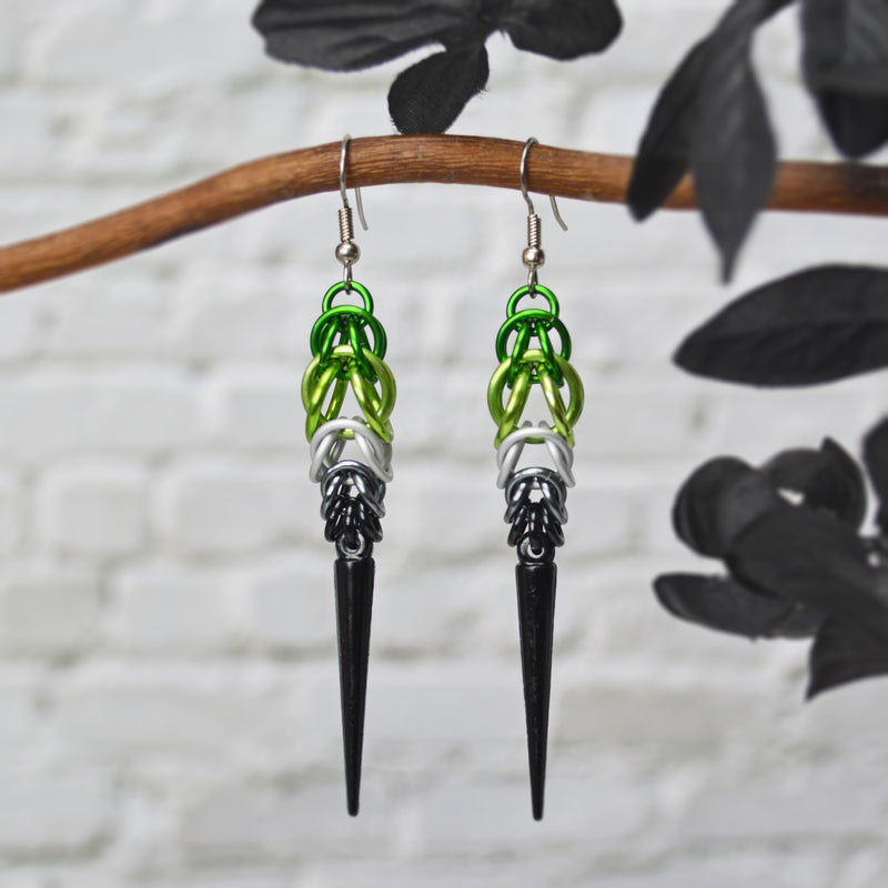 Aromantic pride chainmaille earrings hanging from a branch. Earrings are green and light green at the top, followed by white, grey and black, with a black acrylic spike at the bottom.  