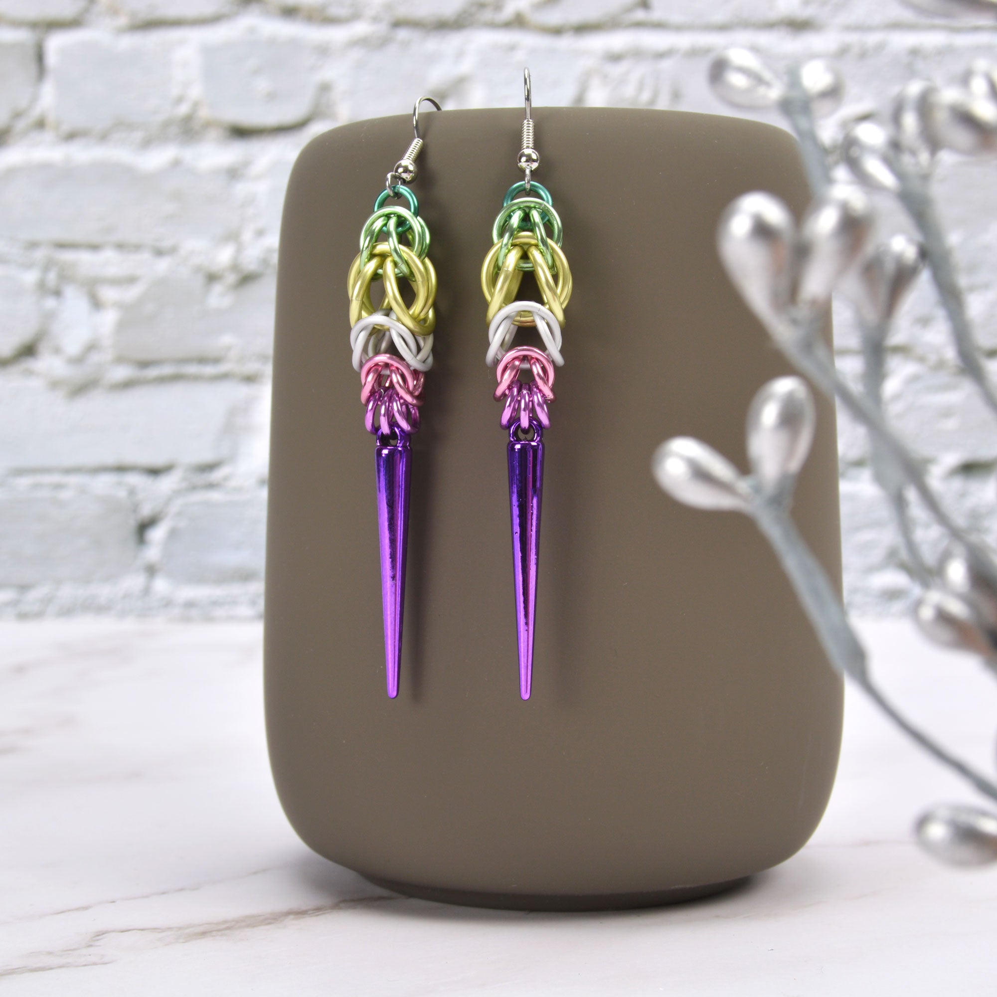 Chainmaille earrings with acrylic spike hanging from a mug. Earrings are in the colors to best match the genderfae pride flag - sage, seafoam, light yellow, white, pink, orchid and violet