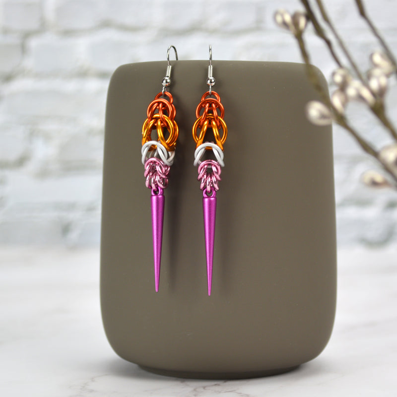 Elegant lesbian pride chainmaille earrings hanging from a dark grey mug. Earrings are dark orange at top, expanding in size to an orange segment, then tapering back down in size in white and pink jump rings. At the bottom, a dark fuchsia spike hangs.
