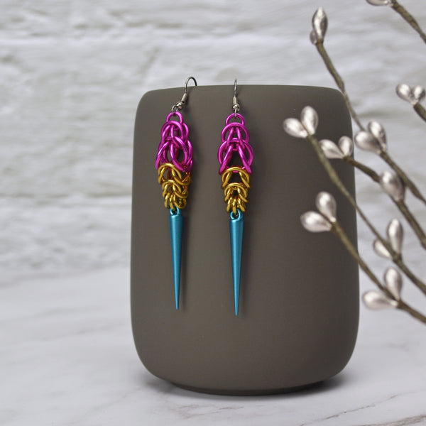 Pansexual Pride chainmaille earrings in vibrant pink, gold and light blue to match the pan pride flag. Earrings are Full Persian chainmail weave at the top, followed by gold and blue Box weave, with a pearlized turquoise spike at the base. The earrings hang from a dark grey mug with a blurred white brick and marble background. 