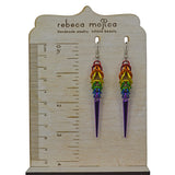Rainbow pride spike earrings hanging from a stand that has inch and centimeter markings on the left side to show the length of the earrings. The bottom tip of the spike earrings reaches about 3.25 inches / 80 mm
