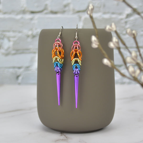 A pair of colorful chainmaille earrings hanging from a grey tumbler mug against a light grey brick wall and light grey marble surface. Earrings are the color of the Xenogenger pride flag: pink at top then peach, orange, pastel yellow, azure, lilac, and violet at the bottom. Chainmaille patter is Full Persian at the top, transitioning to Box Weave, with a long acrylic lilac spike at the base. 