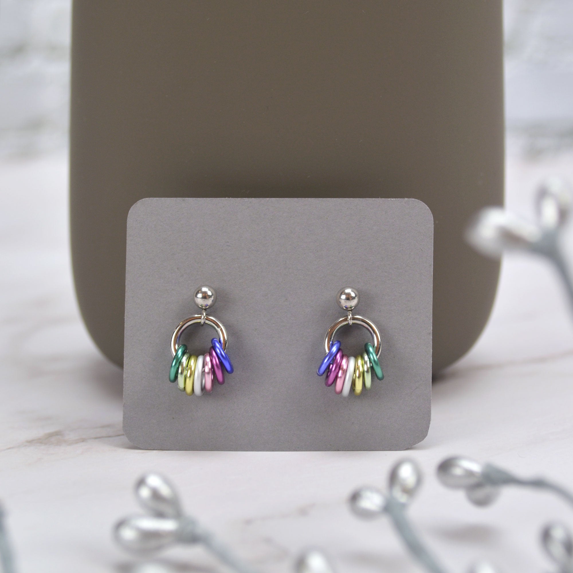 A pair of tiny chainmaille displayed on a small grey card. Earrings have a small stainless steel ball at the top of each post, with a medium size steel ring hanging from the ball. Small anodized aluminum jump rings hang from the medium steel ring in colors to best match the genderfae pride flag: age, seafoam, pastel yellow, white, pink, dusty rose and mystic lavender. The colors appear in mirror image on each earring. Earring card is displayed leaning against the bottom of a darker grey mug.