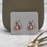 A pair of tiny lesbian pride chainmaille displayed on a small grey card. Earrings have a small stainless steel ball at the top of each post, with a medium size steel ring hanging from the ball. Small anodized aluminum jump rings hang from the medium steel ring in colors to best match the lesbian pride flag: dark orange, orange, white, pink and cranberry. The colors appear in mirror image on each earring. Earring card is displayed leaning against the bottom of a darker grey mug.