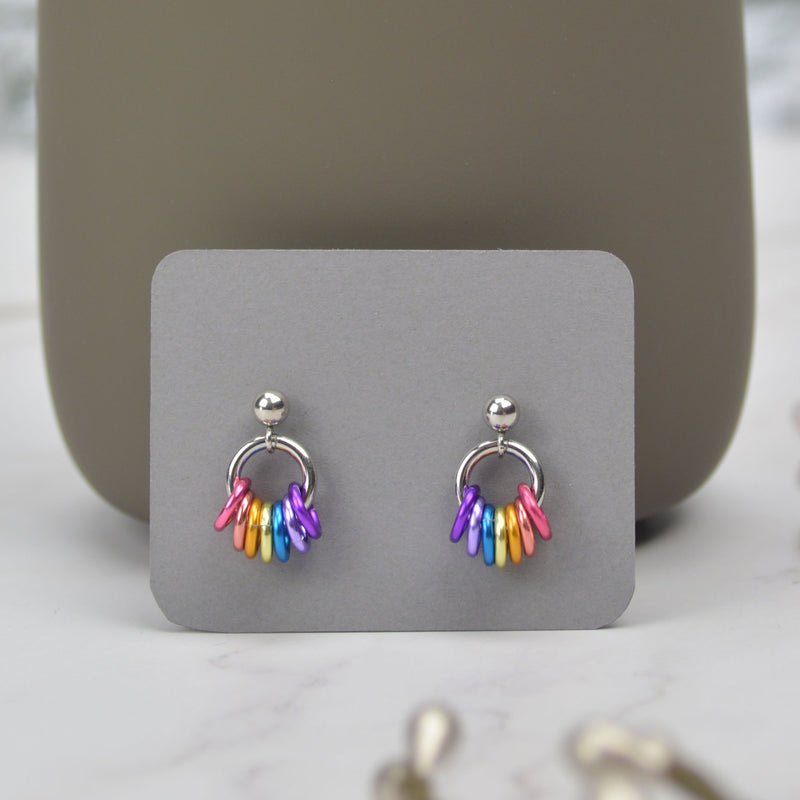 A pair of tiny chainmaille displayed on a small grey card. Earrings have a small stainless steel ball at the top of each post, with a medium size steel ring hanging from the ball. Small anodized aluminum jump rings hang from the medium steel ring in colors to best match the xenogender pride flag: pink, peach, orange, pastel yellow, azure, lilac, and violet. The colors appear in mirror image on each earring. Earring card is displayed leaning against the bottom of a darker grey mug.