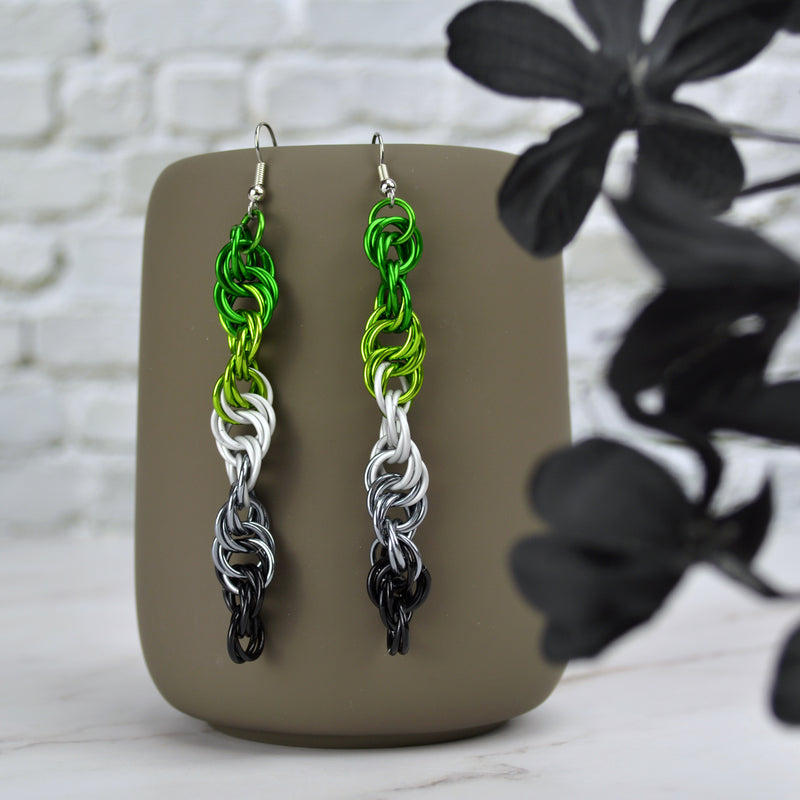 A pair of colorful chainmaille earrings hang from a grey tumbler mug against a light white-grey brick wall and light grey marble surface. Earrings are the color of the Aromantic pride flag: green, lime, white, grey, black. Chainmaille pattern is the Helix weave, which is a long thin spiral. Each earring spirals in an opposite direction