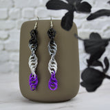 Long and thin chainmaille spiral earrings hang from a grey tumbler against a light grey brick wall and light grey marble surface. Earrings are the color of the asexual pride flag: black, grey, white and violet