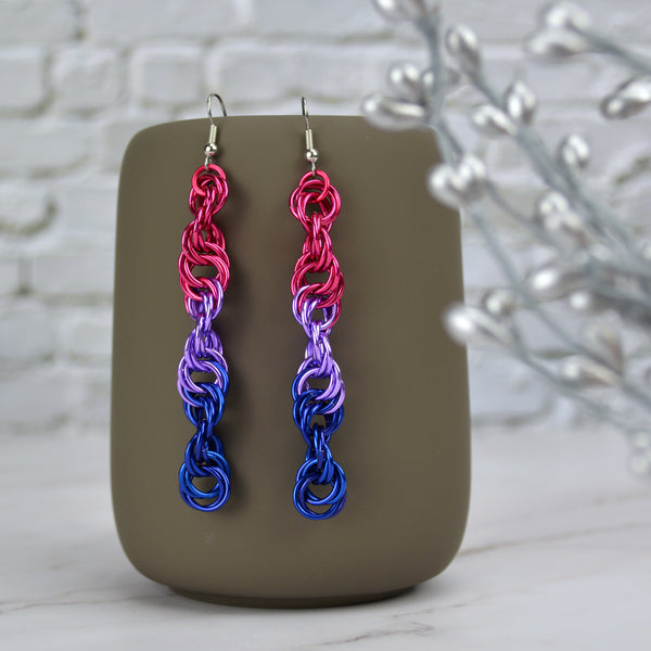 Long and thin chainmaille spiral earrings hang from a grey tumbler against a light grey brick wall and light grey marble surface. Earrings are the color of the bisexual pride flag: pink, lilac, blue