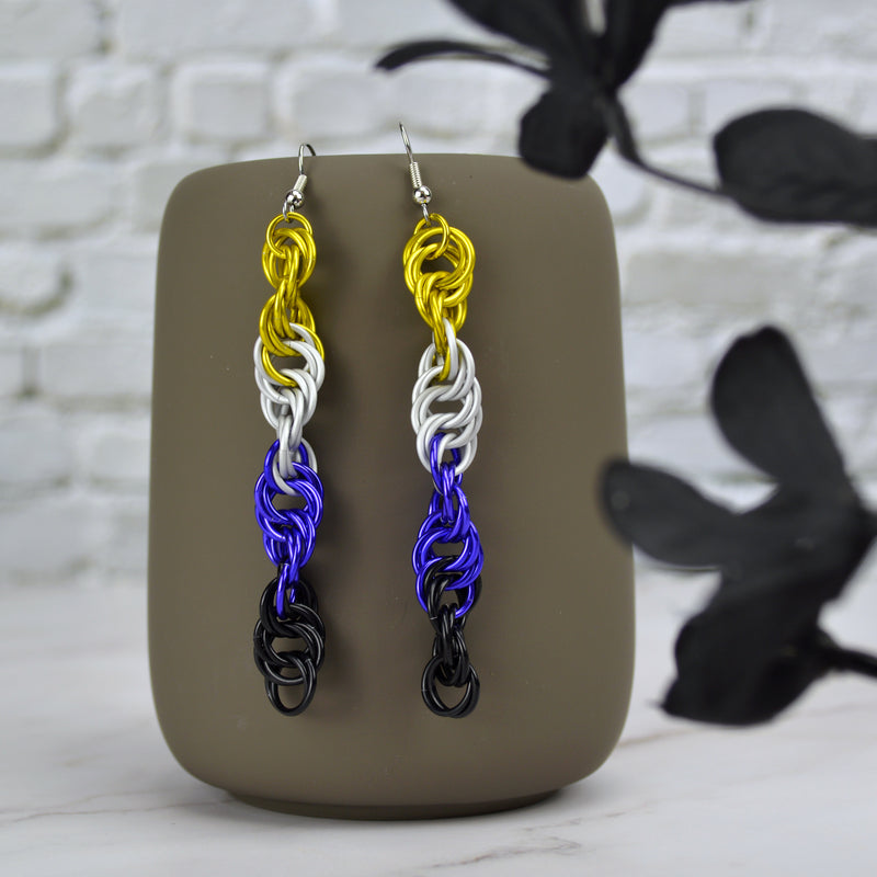 Long and thin chainmaille spiral earrings hang from a grey tumbler against a light grey brick wall and light grey marble surface. Earrings are the color of the nonbinary pride flag: yellow, white, purple and black