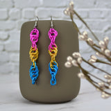 Long and thin chainmaille spiral earrings hang from a grey tumbler against a light grey brick wall and light grey marble surface. Earrings are the color of the pansexual pride flag: pink, gold and blue