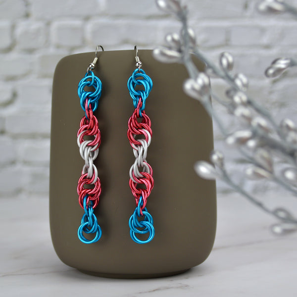 Long and thin chainmaille spiral earrings hang from a grey tumbler against a light grey brick wall and light grey marble surface. Earrings are the color of the transgender pride flag: blue, pink and whit