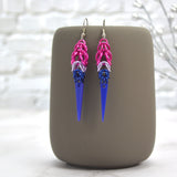 A pair of colorful chainmaille earrings hanging from a grey tumbler mug against a light grey brick wall and light grey marble surface. Earrings are the color of the Bisexual pride flag: pink at top then then lilac, then blue. Chainmaille pattern is Full Persian at the top, transitioning to Box Weave, with a long, flat, laser-cut blue acrylic spike at the base