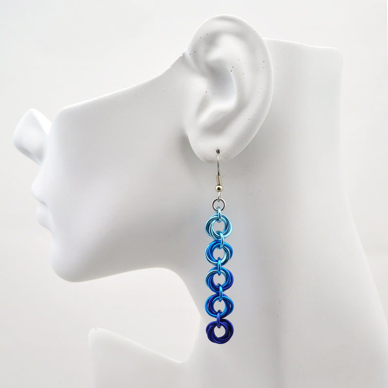 Closeup of chainmaille earrings on display head. Earrings are 5 knots linked vertically and transitioning from a top knot of light blue to a bottom knot of dark blue. Five Knot Earrings by Rebeca Mojica