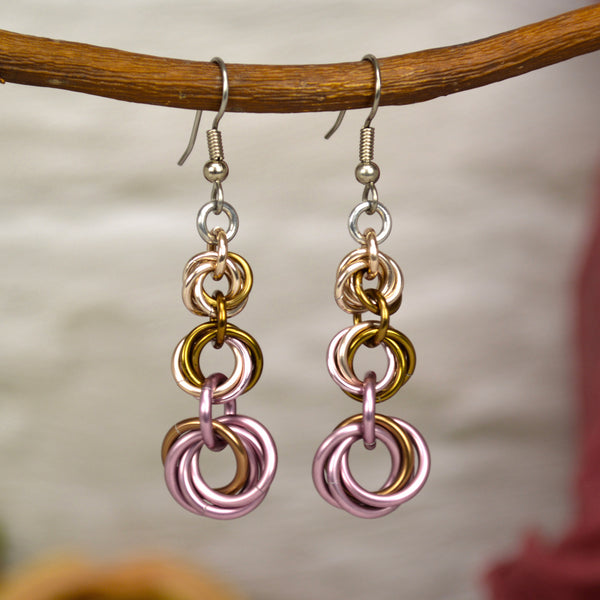 Feminine chain link earrings in rose gold, russet and dusty pink on silver earwire. Displayed hanging from a branch, with a blurry white brick wall background. Each earring has 3 vortices in ascending size. The colors are arranged in an ombre with rose gold and a bit of russet in the top smallest knot, and dusty pink and russet at the base. The middle knot has all 3 colors.