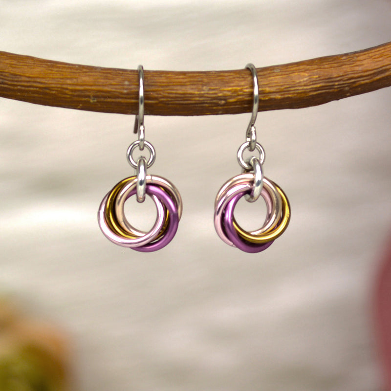 Dainty and feminine chainmaille earrings hanging from a branch. Each earring has a small "knot" of 4 jump rings in rose gold, russet and pink colors on a stainless steel earwire. The background is a blurry off-white color brick wall.