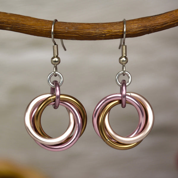 Soft pink and brown chainmaille earrings hanging from a branch. Each earring has a "knot" of 3 large jump rings in rose gold, russet and pink on a stainless steel earwire. The background is a blurry off-white color brick wall.