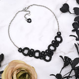 Black chain necklace on a white marble background. The necklace is made of intertwined large and small "knots" of black links, which form an overall V shape for the front half of the necklace. The rear portion is a single strand of thin stainless steel chain. Displayed with black flowers and a beige rose for a gothic look.