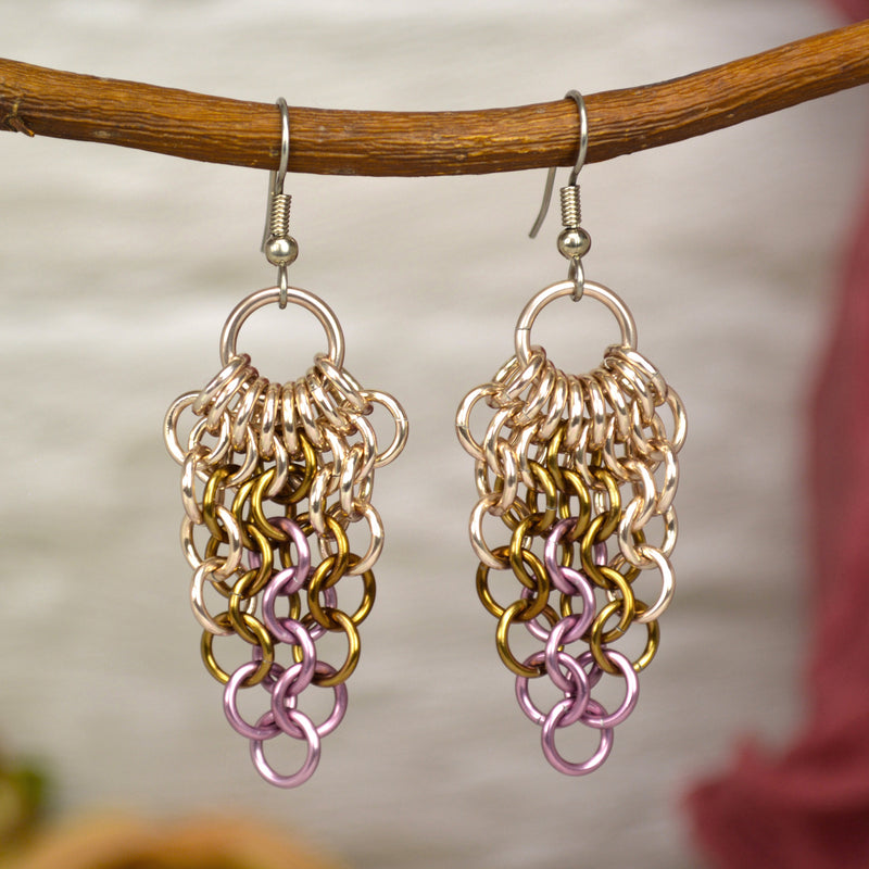 Mesh chainmaille earrings hang from a branch. Each earring has a medium size rose gold color link at the top. Smaller links hang from the top link, first in rose gold, then russet and finally pink at the very bottom. The links form a mesh that tapers to a point, with only a single pink link at the bottom. The background is a blurry off-white color brick wall.