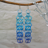 Large link chain mesh earrings hanging from a branch. The long earrings are light blue at the top, azure in the middle and dark blue at the bottom. The background is a blurred light grey brick wall.