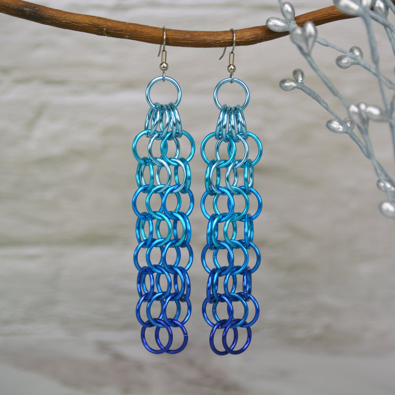 Large link chain mesh earrings hanging from a branch. The long earrings are light blue at the top, azure in the middle and dark blue at the bottom. The background is a blurred light grey brick wall.
