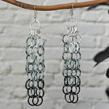 Large link chain mesh earrings hanging from a branch. The long earrings are silver at the top, grey in the middle and black at the bottom tip. The background is a blurred light grey brick wall.