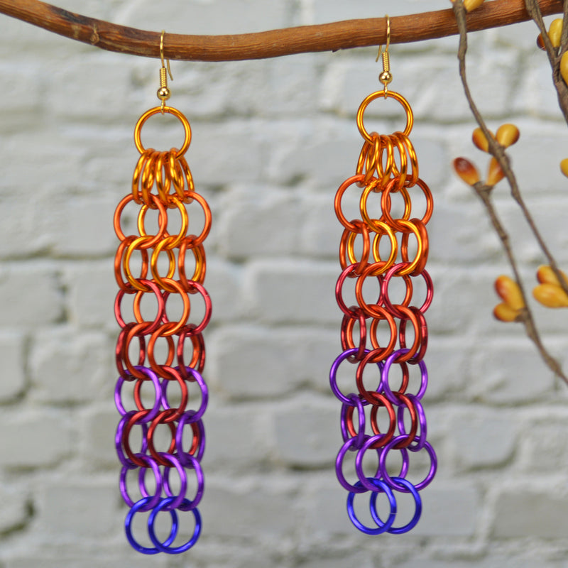 arge link chain mesh earrings hanging from a branch. The long earrings are light bright orange at the top, red in the middle, transitioning to violet and purple at the bottom. The background is a blurred light grey brick wall.