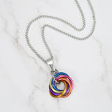Aerial view of a small, circular chainmaille pendant resting against a light grey marble surface. The pendant consists of small interlocking aluminum links (Möbius weave) hanging from a simple, smooth steel pendant bail with a stainless steel chain. The colors of the pendant match the xenogender pride flag: pink, peach, orange, pastel yellow, azure, lilac, and violet. 