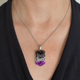 Closeup of a chainmaille pendant in asexual pride colors hanging from a thin steel chain. Pendant is shown worn around the neck of a light-skinned woman wearing a black top with a fluid, V neckline. Pendant is made of four horizontal segments of the chainmail Byzantine pattern. From top to bottom the segments are: black, grey, white and violet.