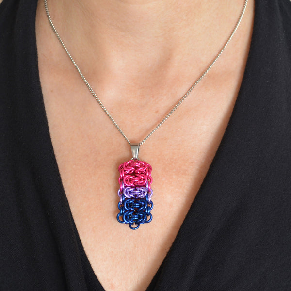 Closeup of a chainmaille pendant in bisexual pride colors hanging from a thin steel chain. Pendant is shown worn around the neck of a light-skinned woman wearing a black top with a fluid, V neckline. Pendant is made of five horizontal segments of the chainmail Byzantine pattern. From top to bottom the segments are: 2 hot pink, 1 lilac and 2 dark blue