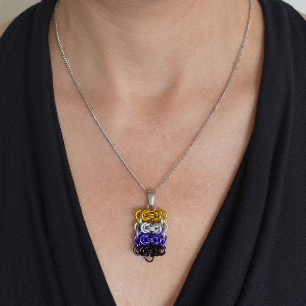Closeup of a chainmaille pendant in nonbinary pride colors hanging from a thin steel chain. Pendant is shown worn around the neck of a light-skinned woman wearing a black top with a fluid, V neckline. Pendant is made of four horizontal segments of the chainmail Byzantine pattern. From top to bottom the segments are: yellow, white, purple and black.