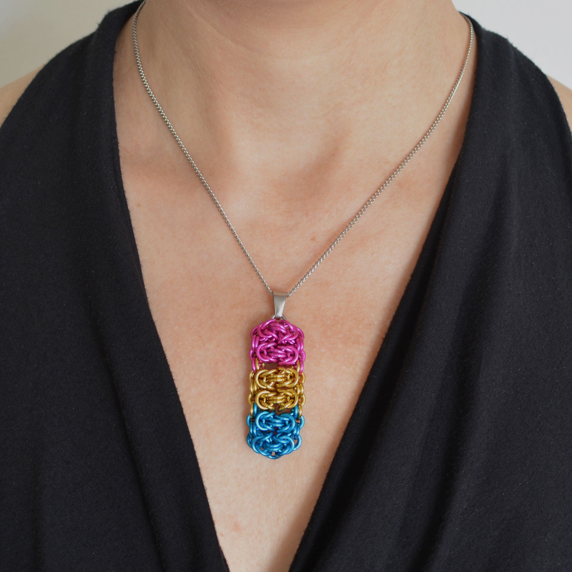 Closeup of a chainmaille pendant in LGBTQ pansexual pride colors hanging from a thin steel chain on a light skinned woman wearing a black top with a fluid, V neckline. Pendant is made of horizontal segments of the Byzantine weave. From top to bottom the segments are: pink; gold and turquoise, two segments in each color.