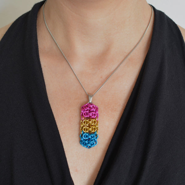 Closeup of a chainmaille pendant in LGBTQ pansexual pride colors hanging from a thin steel chain on a light skinned woman wearing a black top with a fluid, V neckline. Pendant is made of horizontal segments of the Byzantine weave. From top to bottom the segments are: pink; gold and turquoise, two segments in each color.