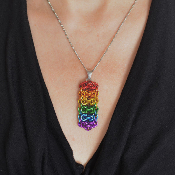 Closeup of a chainmaille pendant in LGBTQ rainbow colors hanging from a thin steel chain on a light skinned woman wearing a black top with a fluid, V neckline. Pendant is made of horizontal segments of the Byzantine weave. Each segment is a different color: red, orange, yellow, green, blue and violet from top to bottom.
