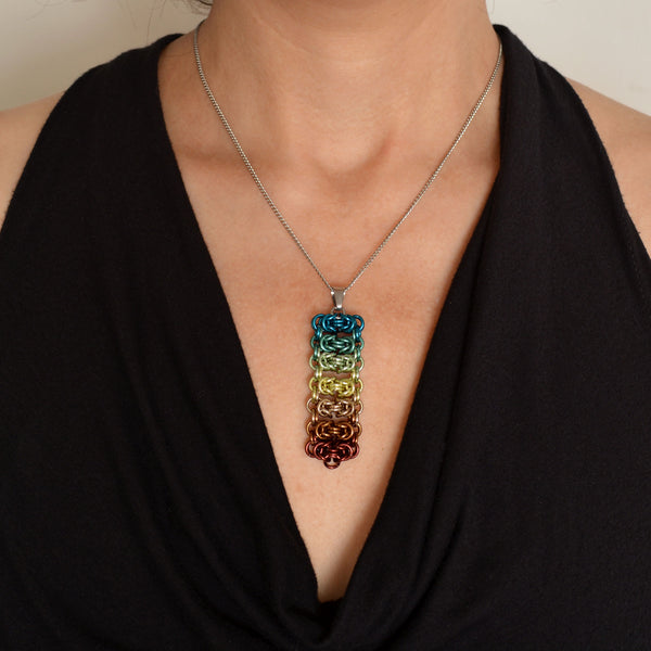 Closeup of a chainmaille pendant in an ombre from teal-green to brown. Pendant is shown worn around the neck of a light-skinned woman wearing a black top with a fluid, V neckline. Pendant is made of seven horizontal segments of the chainmail Byzantine pattern. From top to bottom the segments are: teal, sage, seafoam, pastel yellow, champagne, russet and brown.
