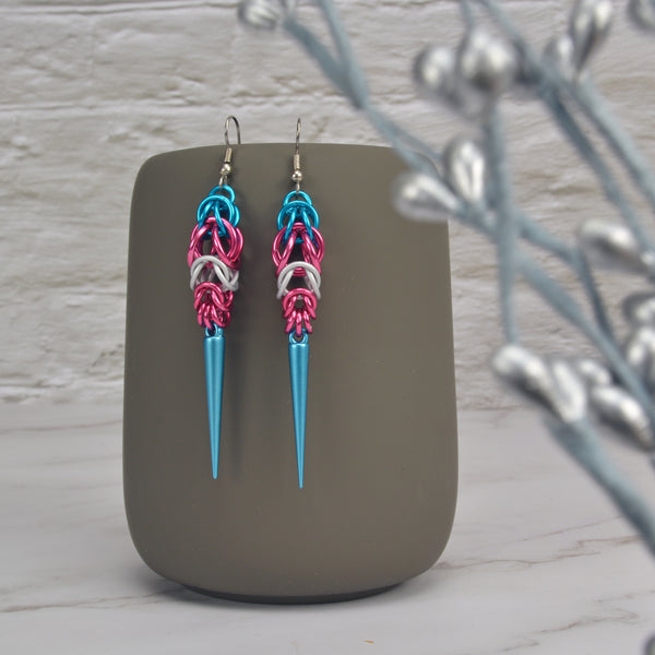 Trans pride chainmaille earrings hanging from a grey tumbler with a white brick background. Earrings are turquoise and pink stripes in the Full Persian weave at the top, followed by white and pink stripes in the Box weave, with a turquoise acrylic spike at the base.