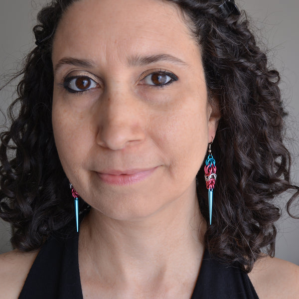 Headshot of chainmaille artisan Rebeca Mojica wearing her Transgender Pride Spike earrings, looking into the camera and smiling.