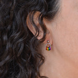 Closeup of a tiny chainmaille earring in LGBTQ rainbow flag colors. Earring is shown on the earlobe of a person with dark brown and grey curly hair. Earring has a small stainless steel ball at the top of the post, with a medium size steel ring hanging from the ball. Small anodized aluminum jump rings hang from the medium steel ring - from left to right the colors are red, orange, yellow, green, blue and violet.