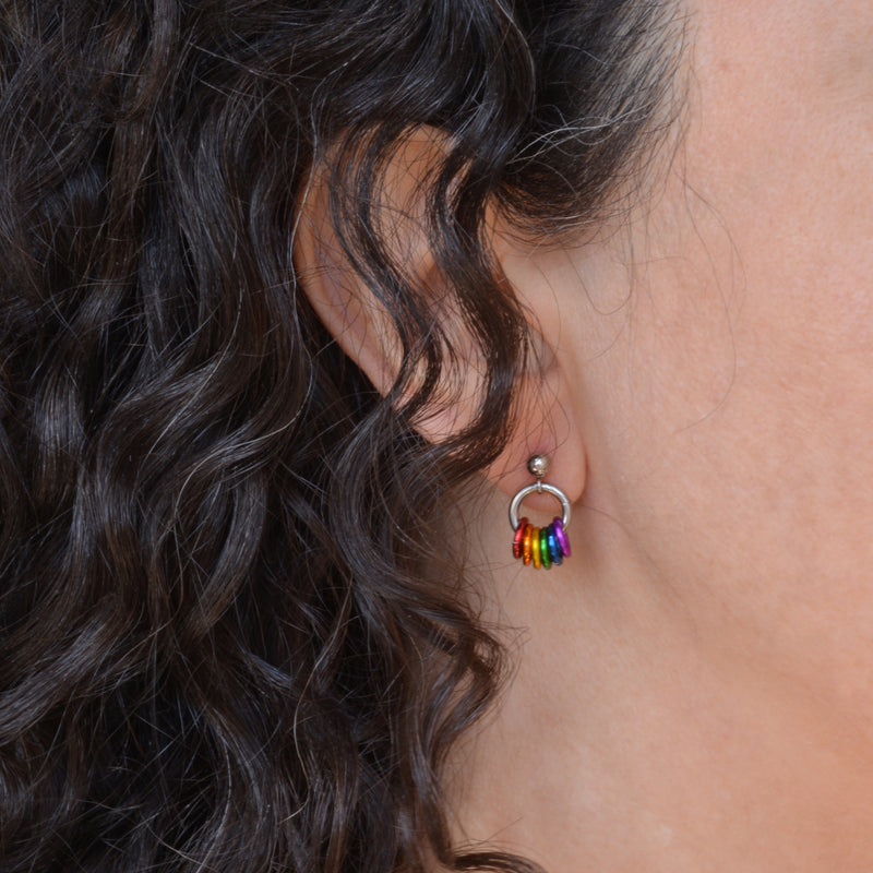 Closeup of a tiny chainmaille earring in LGBTQ rainbow flag colors. Earring is shown on the earlobe of a person with dark brown and grey curly hair. Earring has a small stainless steel ball at the top of the post, with a medium size steel ring hanging from the ball. Small anodized aluminum jump rings hang from the medium steel ring - from left to right the colors are red, orange, yellow, green, blue and violet.