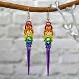 Rainbow pride earrings hanging from a branch. Earrings are red and orange at the top, followed by yellow and green, and then blue, with a long violet spike at the base