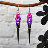 Chainmaille earrings hanging from a branch. Earrings are pink at the top in the Full Persian 6-in-1 weave, transitioning to violet links in box weave, and finishing with small black links and a long black spike at the bottom. The background is a blurred off-white b rick wall.