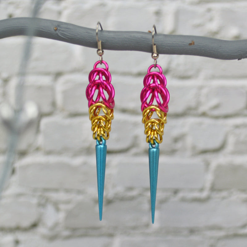 Pansexual pride chainmaille earrings hanging from a branch with a blurred white brick background. Earrings are hot pink at the top in the Full Persian weave, followed by gold links in the Box weave, with a light blue acrylic spike at the base.