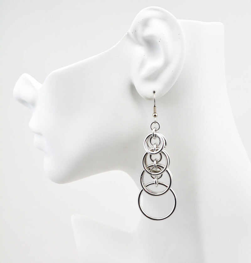 Statement hoop earrings on a white display on a white background. Earrings are 3" long, consisting of stacked intertwined chainmaille jump rings that get larger as you move down the earrings. Display head is shown in profile, facing left.